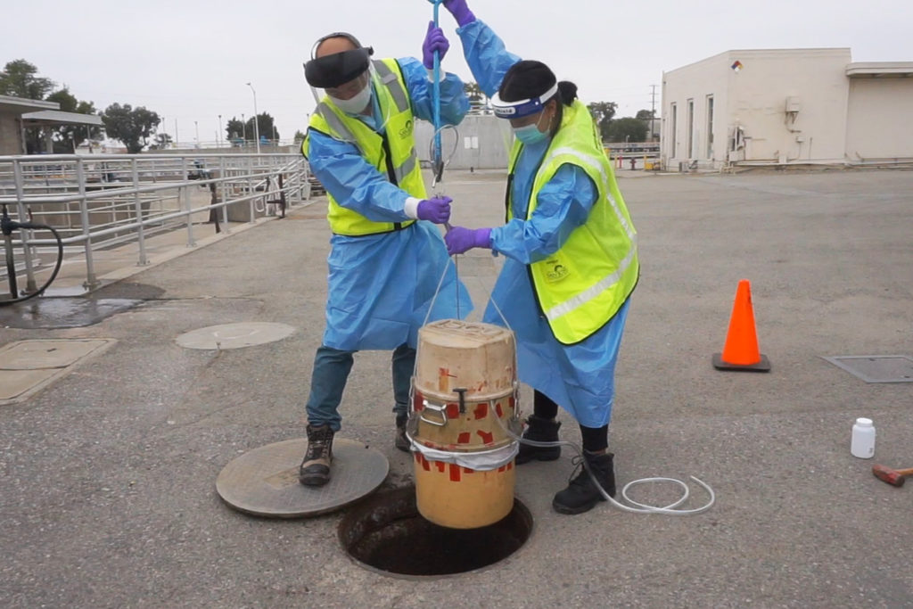 Two people in protective gear holding device over a manhole