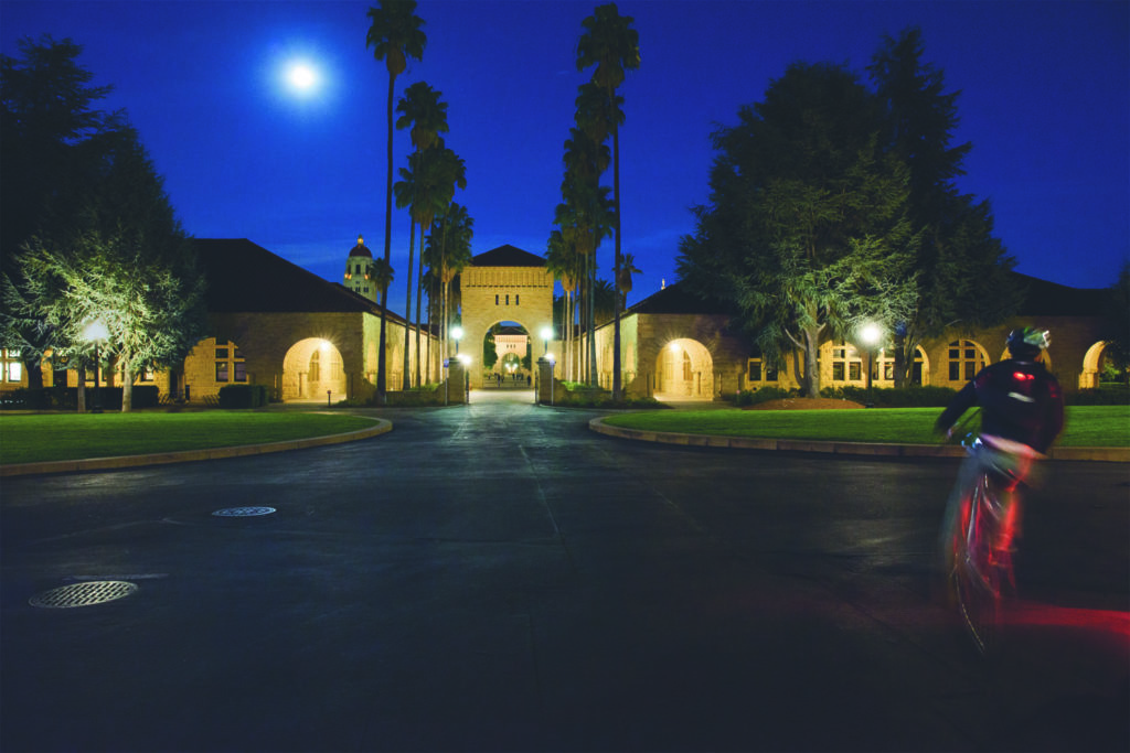 Student biking near the Main Quad at night, with buildings lit by moon and exterior lighting