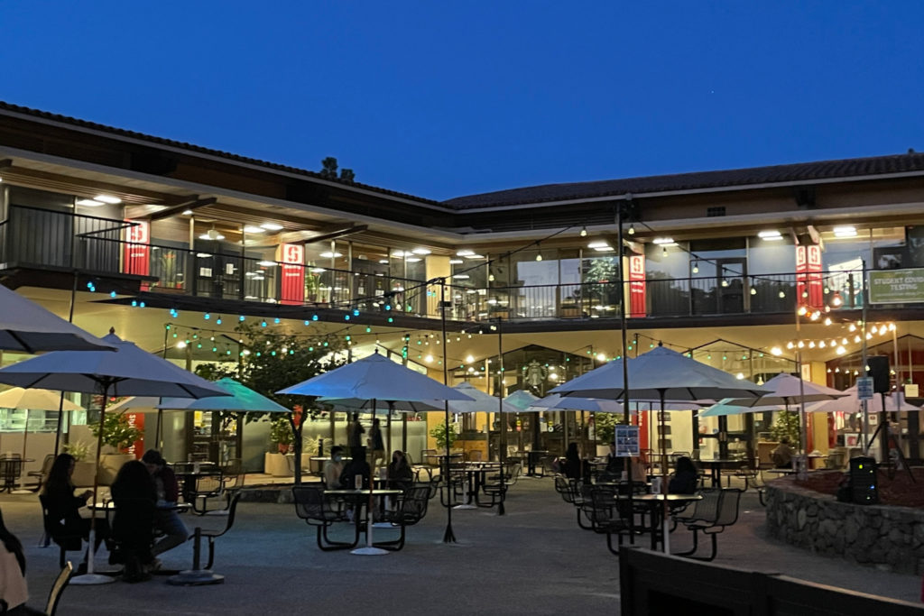 Tresidder exterior, umbrellas, night and string lights, students seated at tables