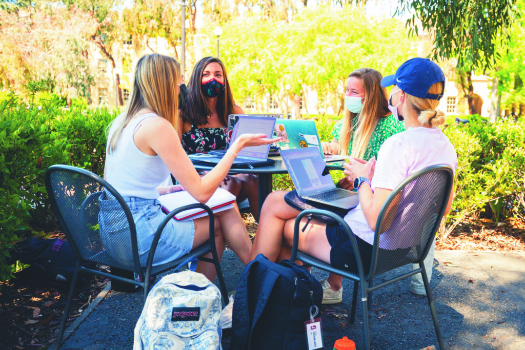 Four students seated around a table outdoors, with face coverings, in conversation