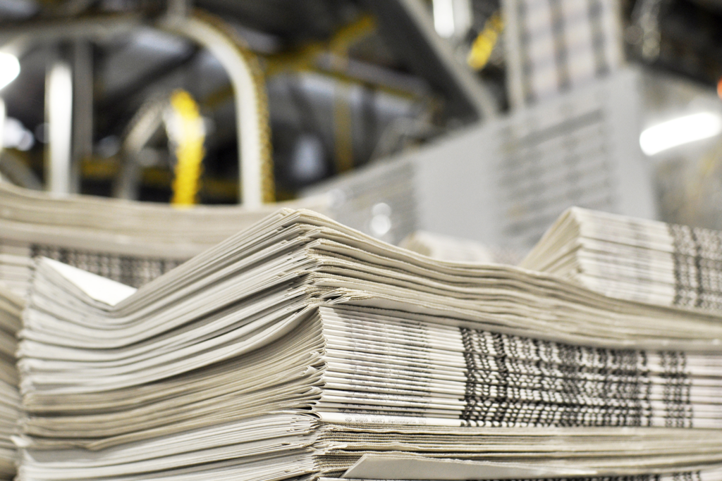 A stack of freshly printed newspapers, in the background printing machines and technical equipment.