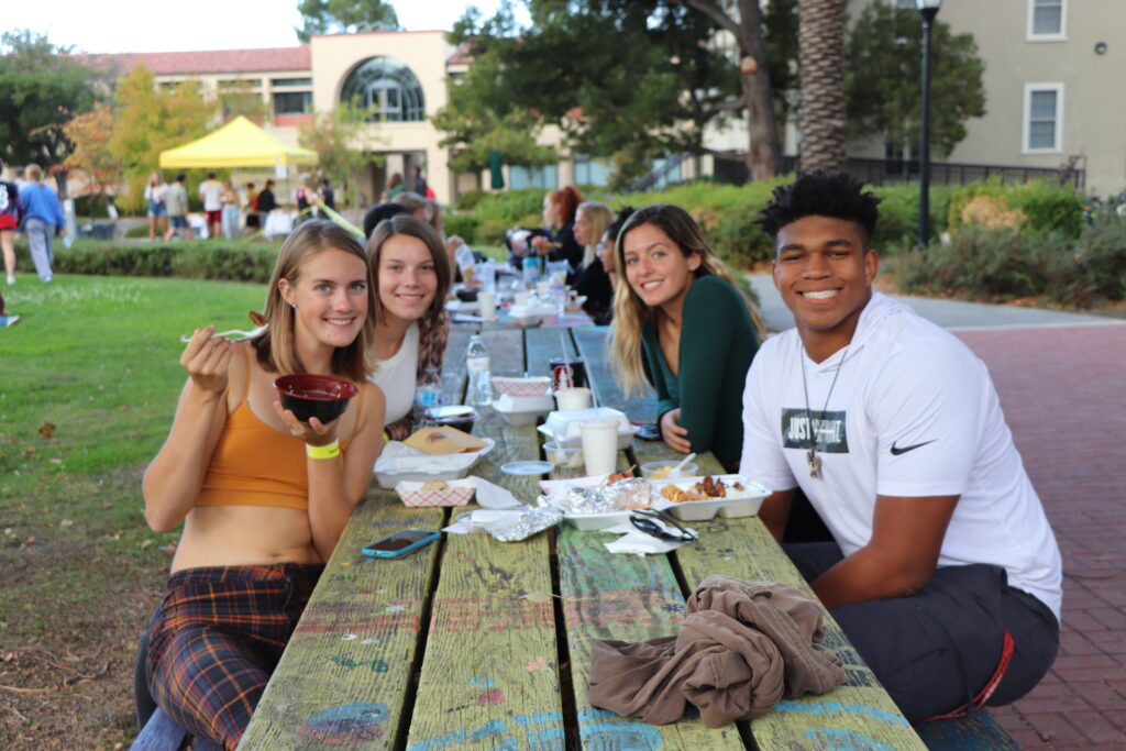 Shows how students gather at a Farm Day. Four students seated at a picnic table eating takeout food.