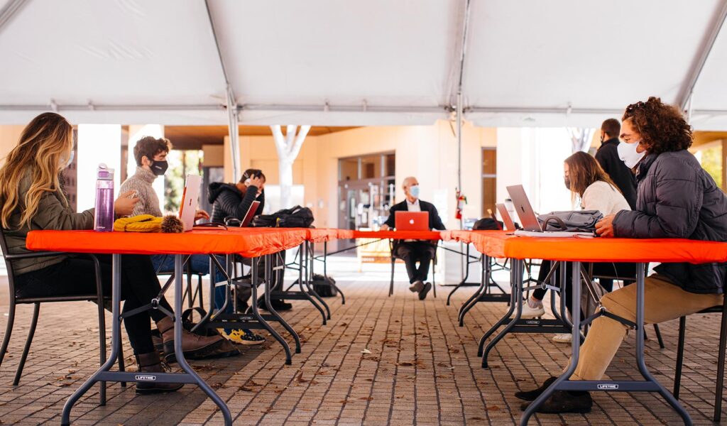 Students and a professor sitting at makeshift desks under a large outdoor tent for outdoor instruction.