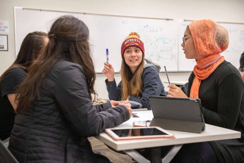 Four students sitting at a desk, near a whiteboard, having a group discussion.