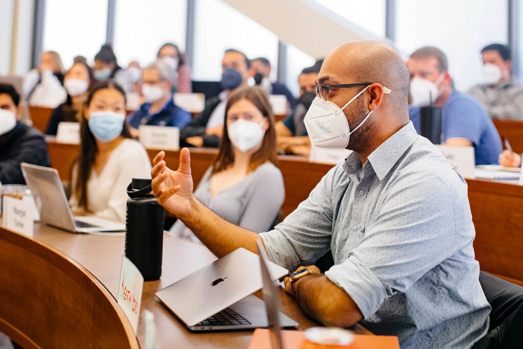 GSB students in a classroom wearing masks, watching intently as a male student holds his hand forward while speaking to the class.