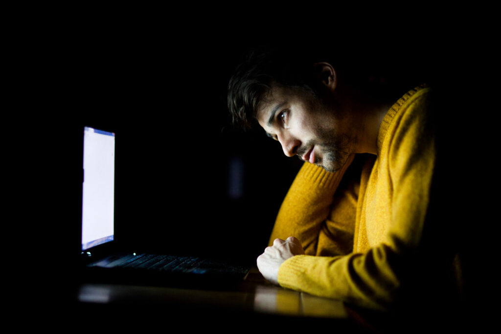 A man wearing a yellow sweater sitting in a dark room, staring blankly into his computer screen.