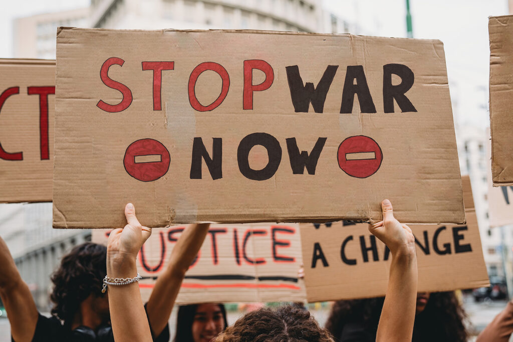 People holding protest signs on a cloudy day in an unknown city. The foremost sign says "stop war now."