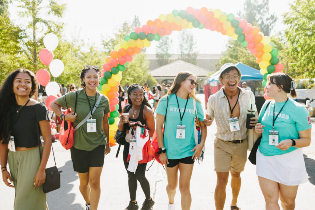 Six students walk through balloon arch, including two in teal t-shirts, outdoors at White Plaza.