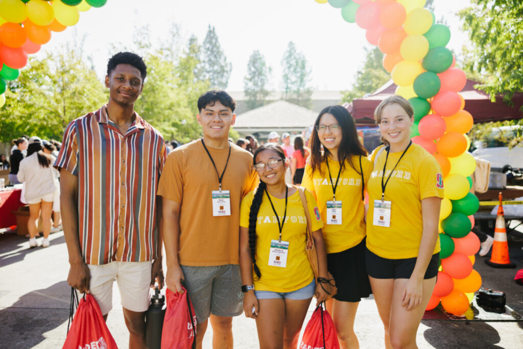 Five students in yellow t-shirts under a balloon arch, holding red backpacks, outside at White Plaza.