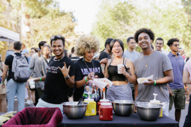 Four students holding hot dogs and standing in front of containers of ketchup and mustard.