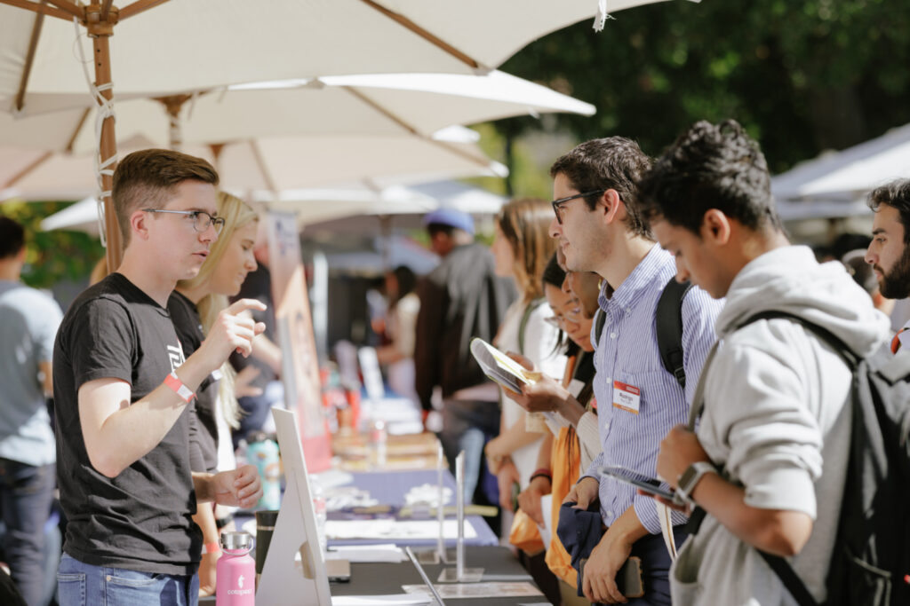 Shows outdoor career fair with long row of tables, employers on one side, students on the other side, getting to know each other