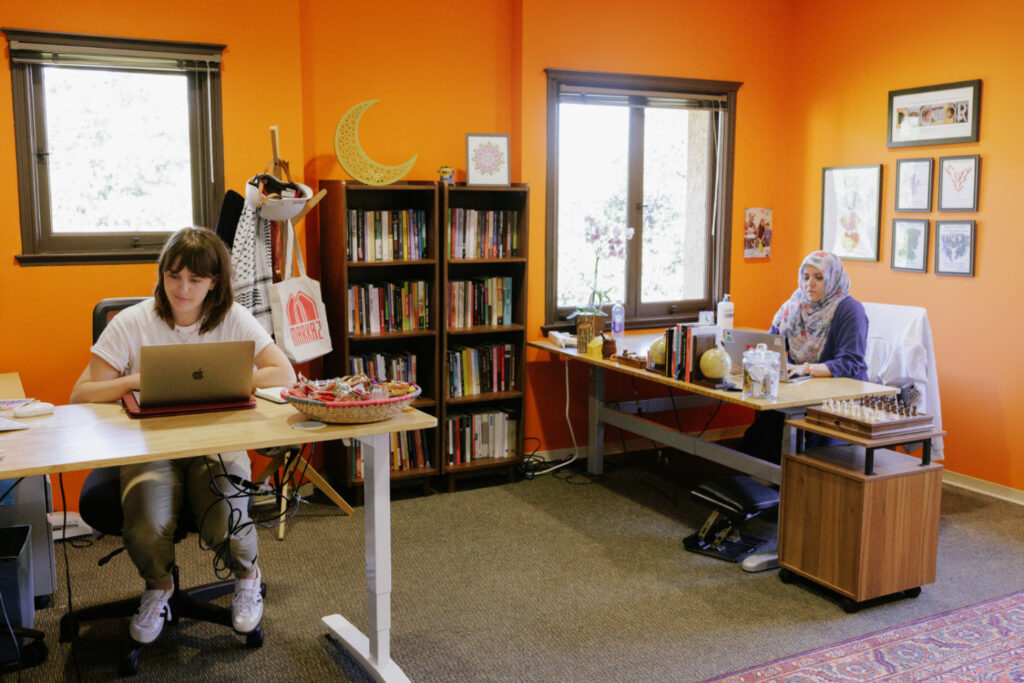 Two staff members sitting at desks inside an office with bookshelves and orange-tinted walls.