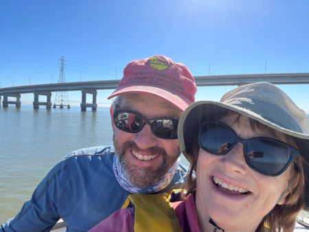 Two people in hats and sunglasses pose in front of a bridge