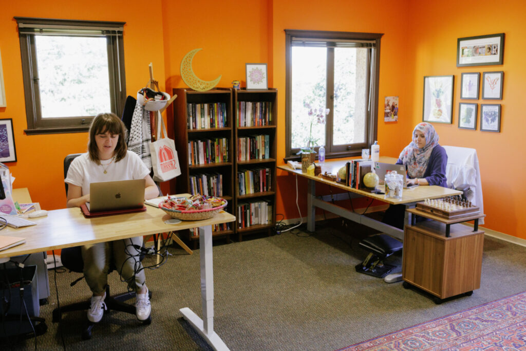 Shows the interior of the Markaz at Stanford, with two staff members seated at desks and decor including a warm orange wall.