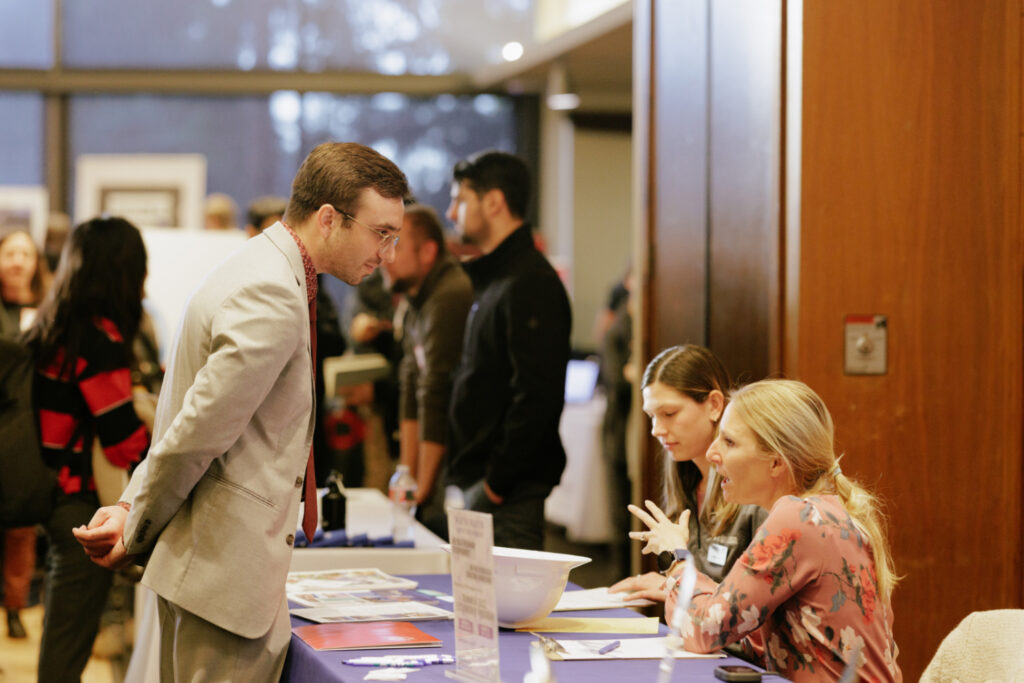 Student wearing a suit and tie, leaning over a table to listen to two people sitting behind the table, which is covered with brochures.