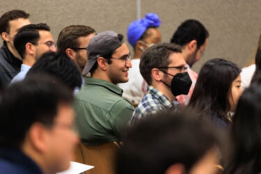 Image shows crowded meeting room and focuses on one conference participant looking up deep in thought while listening to a speaker.