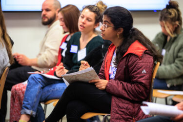 Image shows crowded meeting room and focuses on one conference participant looking up deep in thought while listening to a speaker.