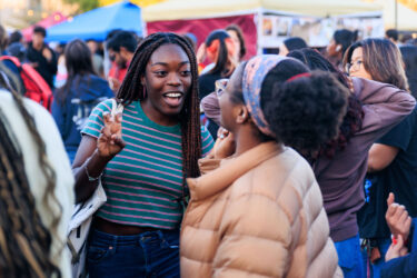 Two friends smiling in the foreground, festival booths in the background.