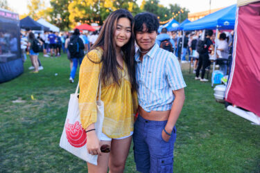 Two friends smiling in the foreground, festival booths in the background.