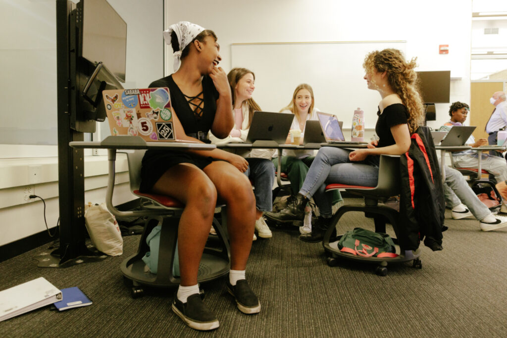 Student seated on a chair on wheels, with a tray below holding a backpack.