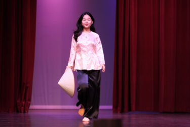 Model wearing pink top, black slacks and carrying a hat.