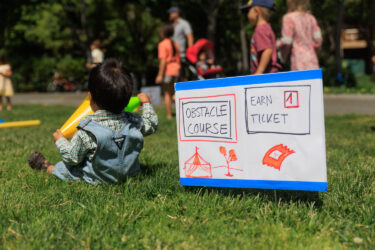 Toddler sitting on grass near an obstacle course.