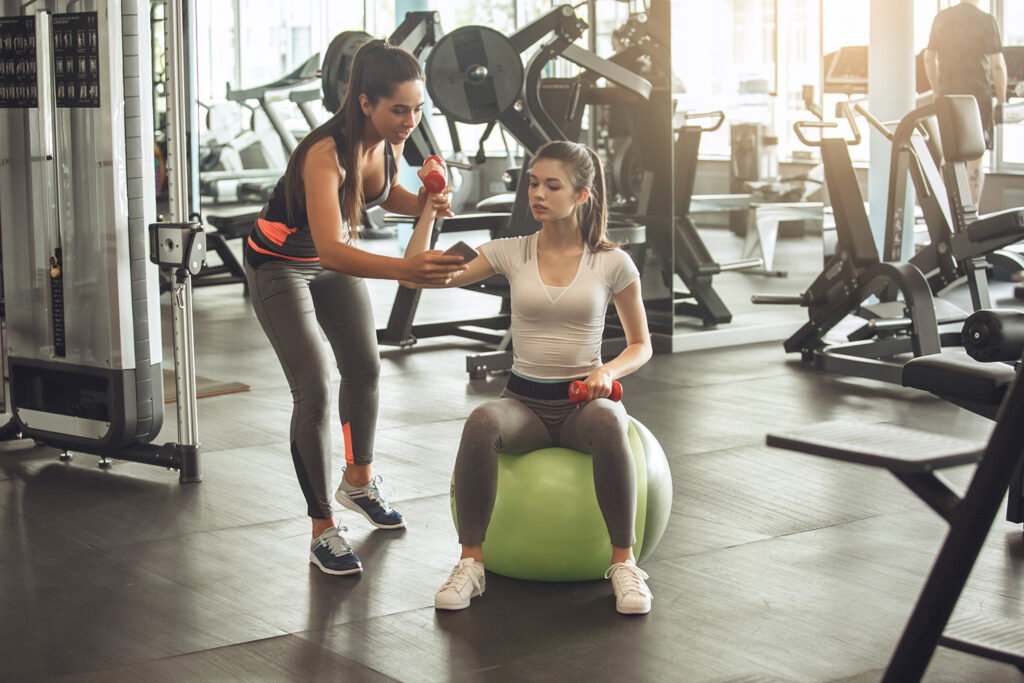 Two women in a gym, one is a trainer looking at her phone, and the other is sitting on an exercise ball.