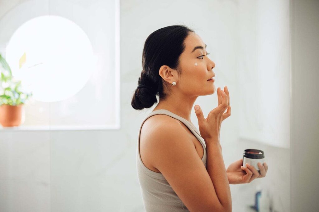 Woman holding face cream and applying it to her cheeks and face.