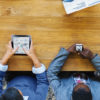 overhead view of people sitting around a table using various phones, tablets, etc.