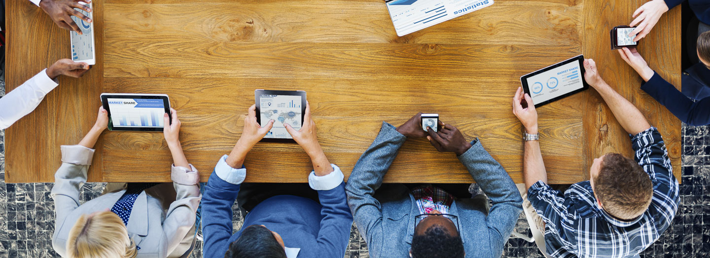 overhead view of people sitting around a table using various phones, tablets, etc.