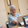 Young man talking with an older woman who sits in front of a computer screen. Logo for the project on another screen.