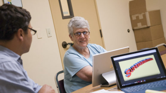 Young man talking with an older woman who sits in front of a computer screen. Logo for the project on another screen.