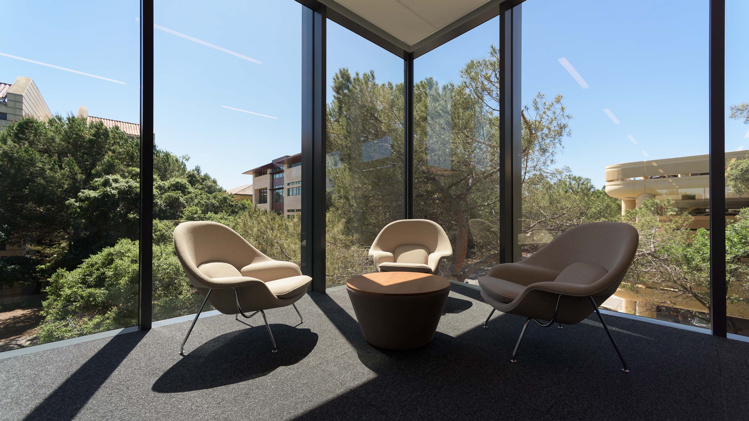 Three empty armchairs surround a small, low round table in a corner of the library with floor-to-ceiling glass windows, with views of treetops outside. (Image credit: L.A. Cicero)