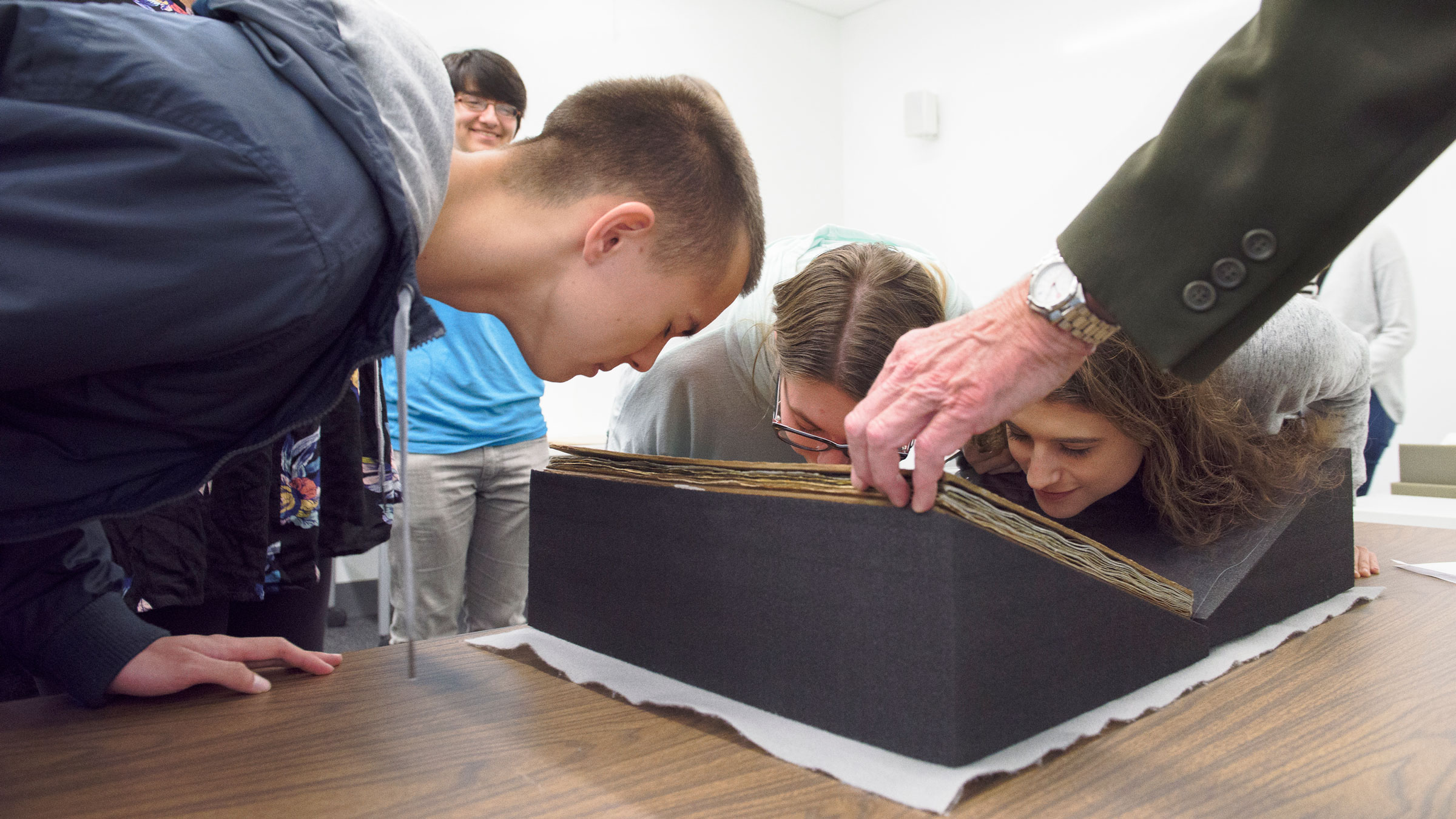 Three undergraduate students, eyes closed, lean close to the cover of a book resting in a V-shaped black foam block resting on a table, to smell the spices infused in its pages. (Image credit: L.A. Cicero)