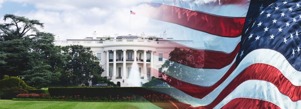 A front-lawn view of the white house, with an american flag flying on the foreground