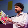 Manu Pakrash holds an origami-stype paper microscope on stage at TED