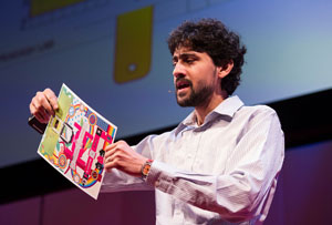 Manu Pakrash holds an origami-stype paper microscope on stage at TED