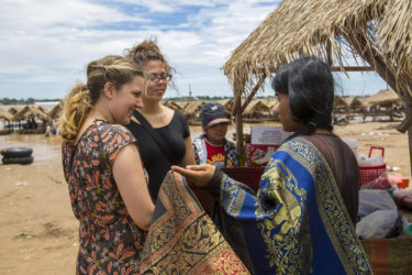 young woman purchasing ornately embroidered fabric at thatched roof stall in an open air market