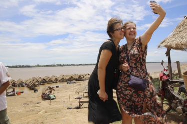 two women taking a selfie in front of thatched roof huts and the Mekong river