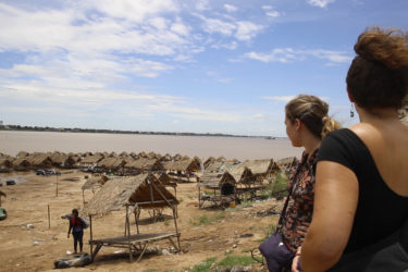 two women look out over thatched roof huts towards the Mekong River