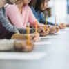 Cropped view of a multiracial group of young men and women sitting in a row at a table, writing with pencils on paper taking a test.