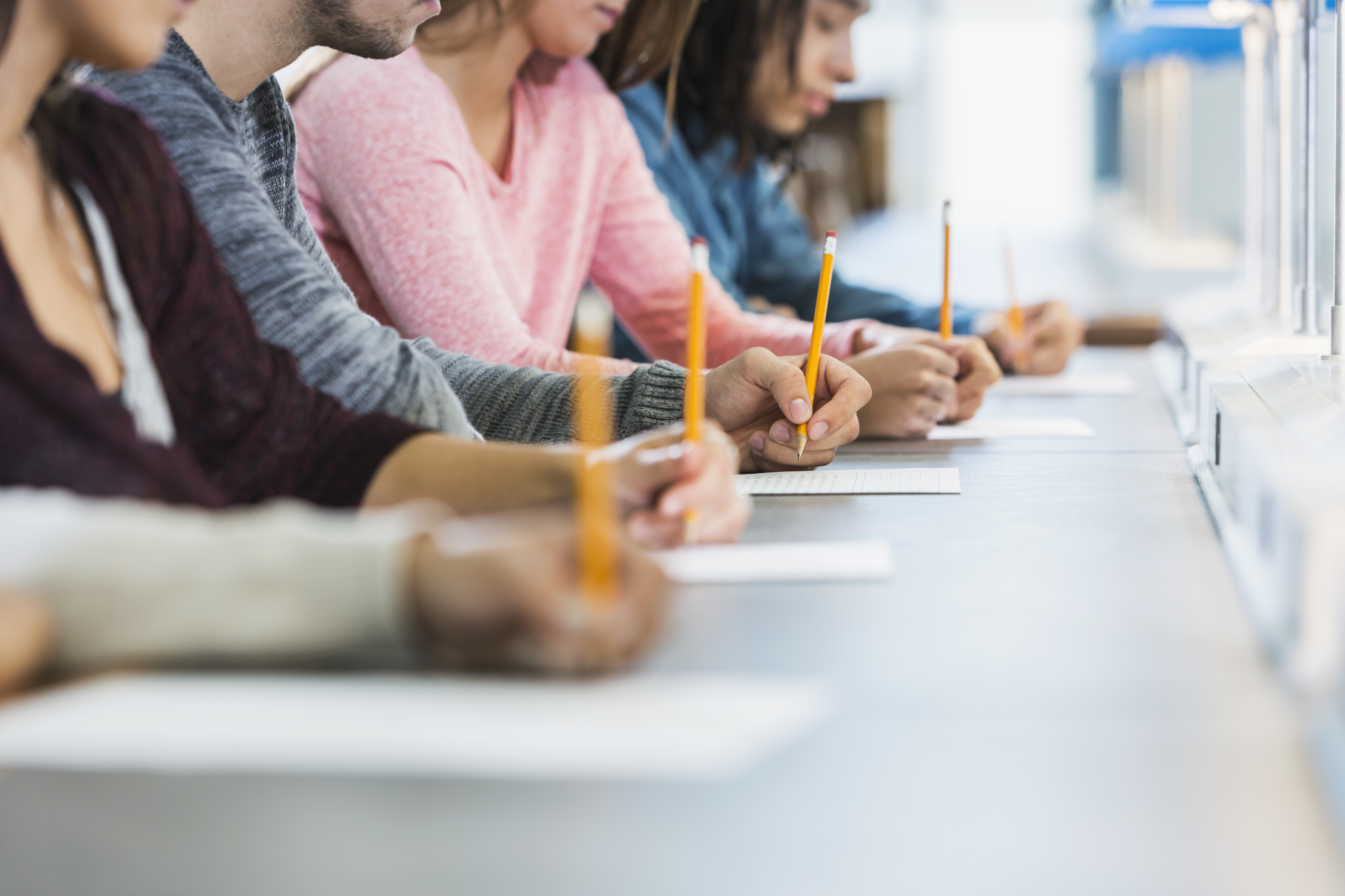 Cropped view of a multiracial group of young men and women sitting in a row at a table, writing with pencils on paper taking a test.