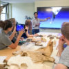 Krish Seetah, assistant professor of anthropology, in the classroom teaching a class on zooarchaeology. Bones are on the table in front of students and the professor is pointing at a 3-D image on a large screen in the front of the class.