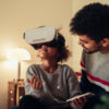 father sitting next to daughter and looking on as she wears a VR headset