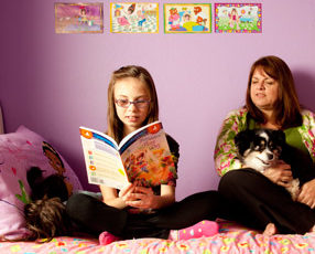 Janet Cartwright sits on a bed with a dog while her daughter reads