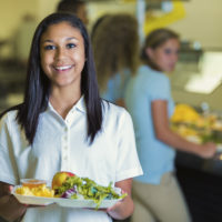 High school student holds a plate of healthy food in the school cafeteria. Her classmates are waiting in line in the background.