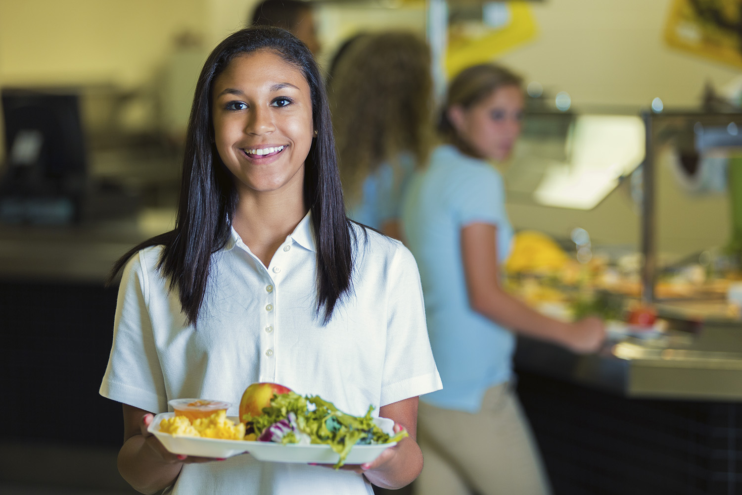 High school student holds a plate of healthy food in the school cafeteria. Her classmates are waiting in line in the background.