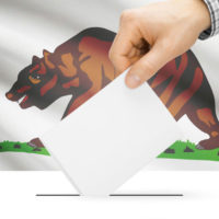 hand inserting ballot into box; California flag in background