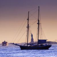 A recreational sailboat in front of the Statue of Liberty, with signs of industry in the background.