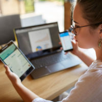 Woman holding phone in one hand, tablet in another, at a laptop computer.